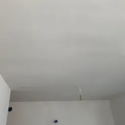 Wnętrze w trakcie gładzenia ścian i sufitu, widoczne przygotowane kable elektryczne wystające z sufitu, ściany pokryte gładzią, okno w prawym dolnym rogu.