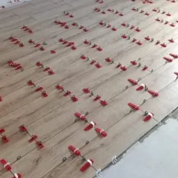 Układanie podłogi z paneli imitujących drewno, z użyciem czerwonych klipsów poziomujących, widoczne gniazdko elektryczne na ścianie w rogu pomieszczenia.