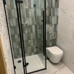 Nowoczesna kabina prysznicowa z czarnymi profilami, biały brodzik, wisząca toaleta i płytki w odcieniach szarości, błękitu i brązu, oraz białe płytki z falistą strukturą na ścianie.