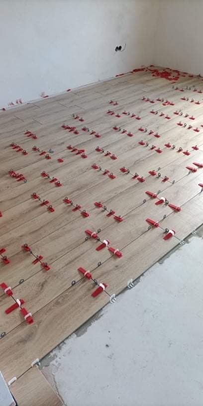 Układanie podłogi z paneli imitujących drewno, z użyciem czerwonych klipsów poziomujących, widoczne gniazdko elektryczne na ścianie w rogu pomieszczenia.