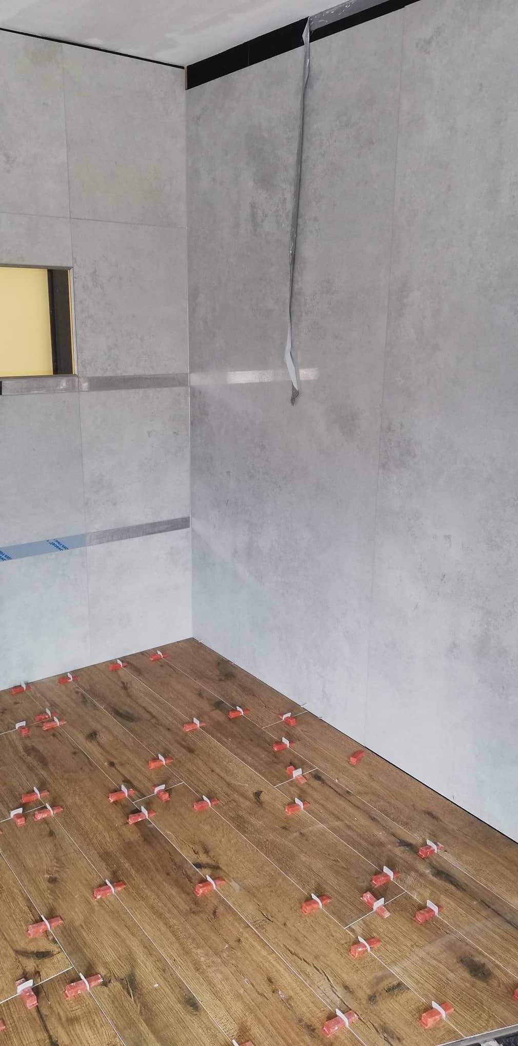 Proces układania drewnopodobnych płytek podłogowych w łazience, z widocznymi krzyżykami dystansowymi i fragmentem ściany wyłożonej szarymi płytkami.