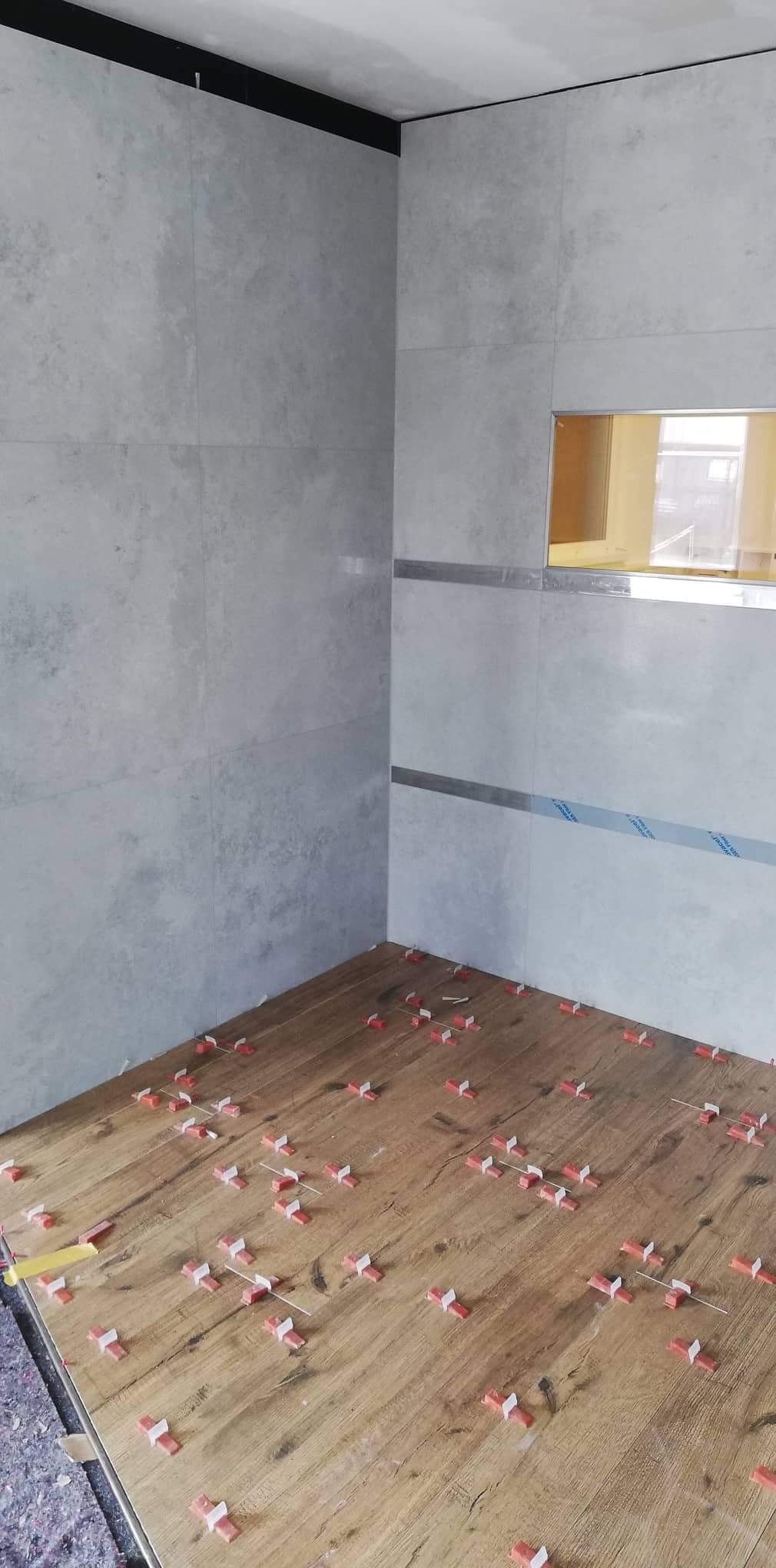 Łazienka w trakcie wykańczania: szare płytki na ścianach z poziomymi listwami dekoracyjnymi i wnęką, drewnopodobne płytki podłogowe z czerwono-białymi krzyżykami dystansowymi.