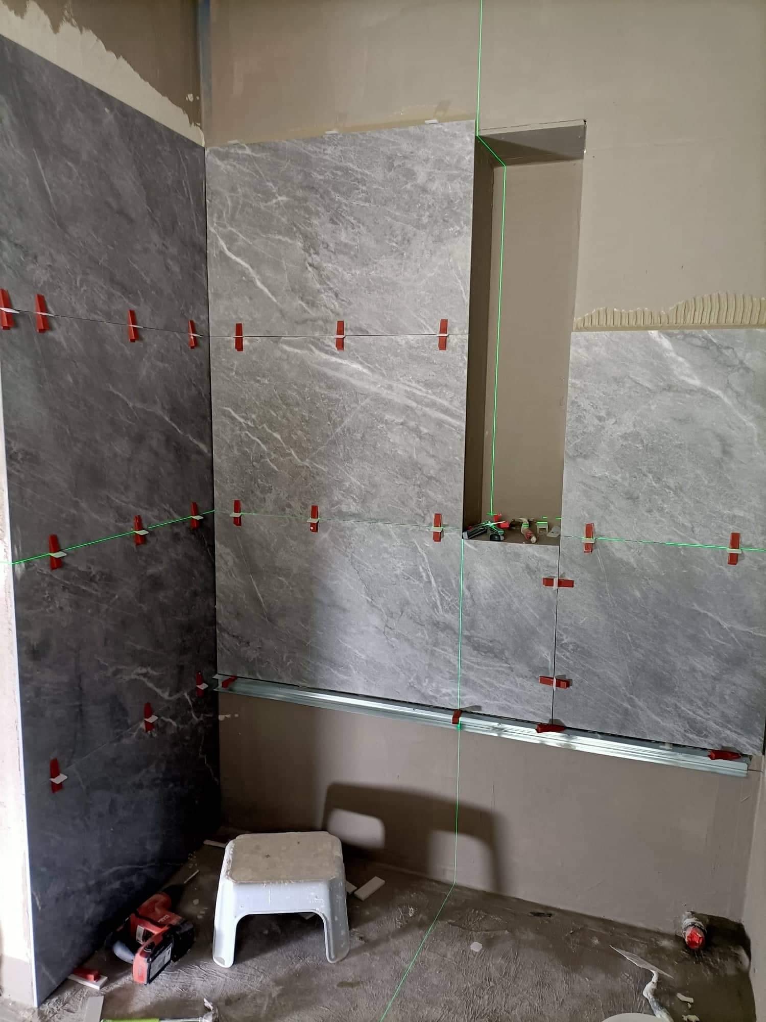 Układanie wielkoformatowych szarych płytek imitujących marmur w łazience, widoczne poziomice laserowe, system poziomowania płytek na ścianach z wnęką, narzędzia glazurnicze i plastikowy stołek...