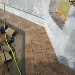 BrukDom Ihor Havryliuk - Układanie brązowej kostki brukowej w Grodzisku Mazowieckim, widoczny wąż ogrodowy i fragment stopy pracownika podczas mycia ułożonej nawierzchni.