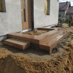 BrukDom Ihor Havryliuk - Wykończone schody wejściowe z brązowej kostki granitowej, częściowo mokre, w trakcie prac ziemnych przy elewacji domu jednorodzinnego.