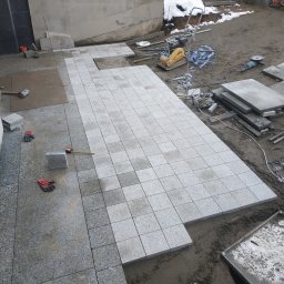 BrukDom Ihor Havryliuk - Układanie jasnoszarej kostki na placu budowy, widoczna ubijarka, szlifierka kątowa, palety z kostką w tle i resztki śniegu.