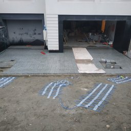 BrukDom Ihor Havryliuk - Montaż ogrzewania podjazdowego z kostki granitowej: widoczne maty grzewcze na ziemi przed częściowo ułożonym podjazdem z kostki granitowej w kolorze szarym, garaż w tle.