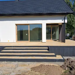 Nowoczesny taras z kostki brukowej w odcieniach beżu i szarości, prowadzący do domu z dużymi oknami przesuwnymi. Widoczne schody i fragment trawnika.