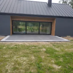 Nowoczesny dom z szarą elewacją i dużymi przesuwnymi drzwiami tarasowymi. Na pierwszym planie szary taras z płytek, myty wodą z węża przez mężczyznę bez koszulki. Widoczne elementy ogrodu.