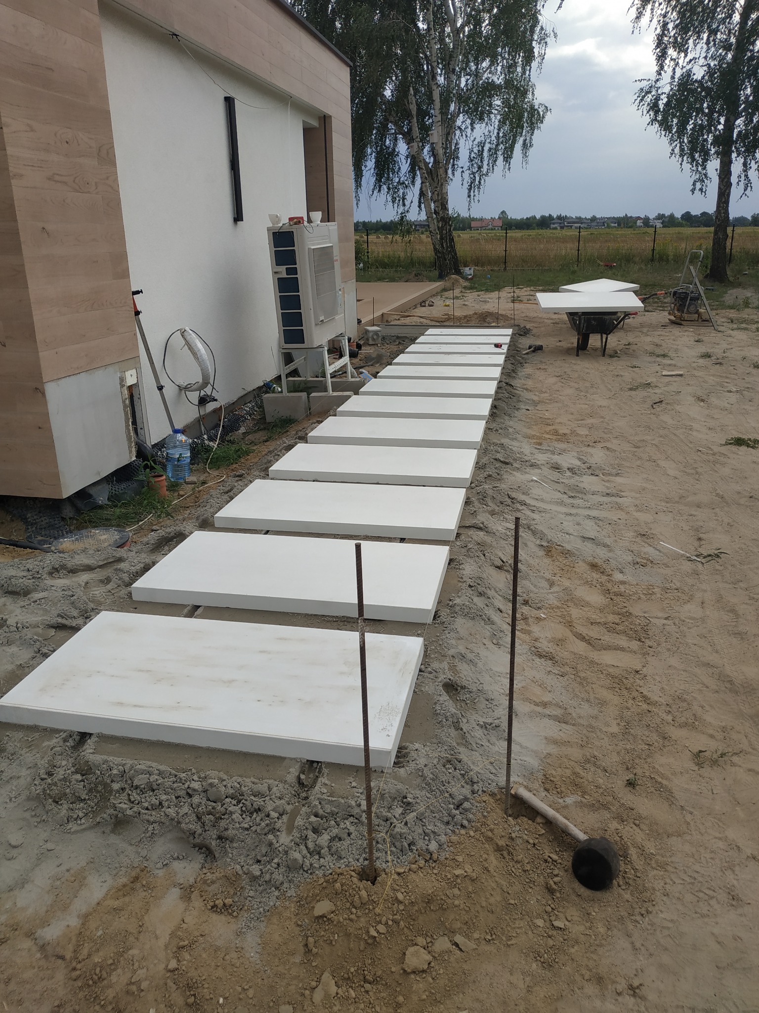 Ułożone białe płyty chodnikowe tworzące ścieżkę wzdłuż budynku, widoczne przygotowane podłoże, narzędzia i taczka w tle.