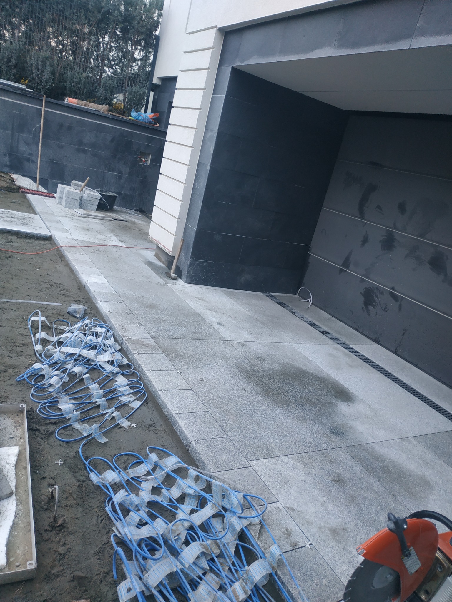 Fragment układania szarej kostki granitowej na podjeździe z widocznymi przewodami ogrzewania podjazdowego i przecinarką do kamienia.