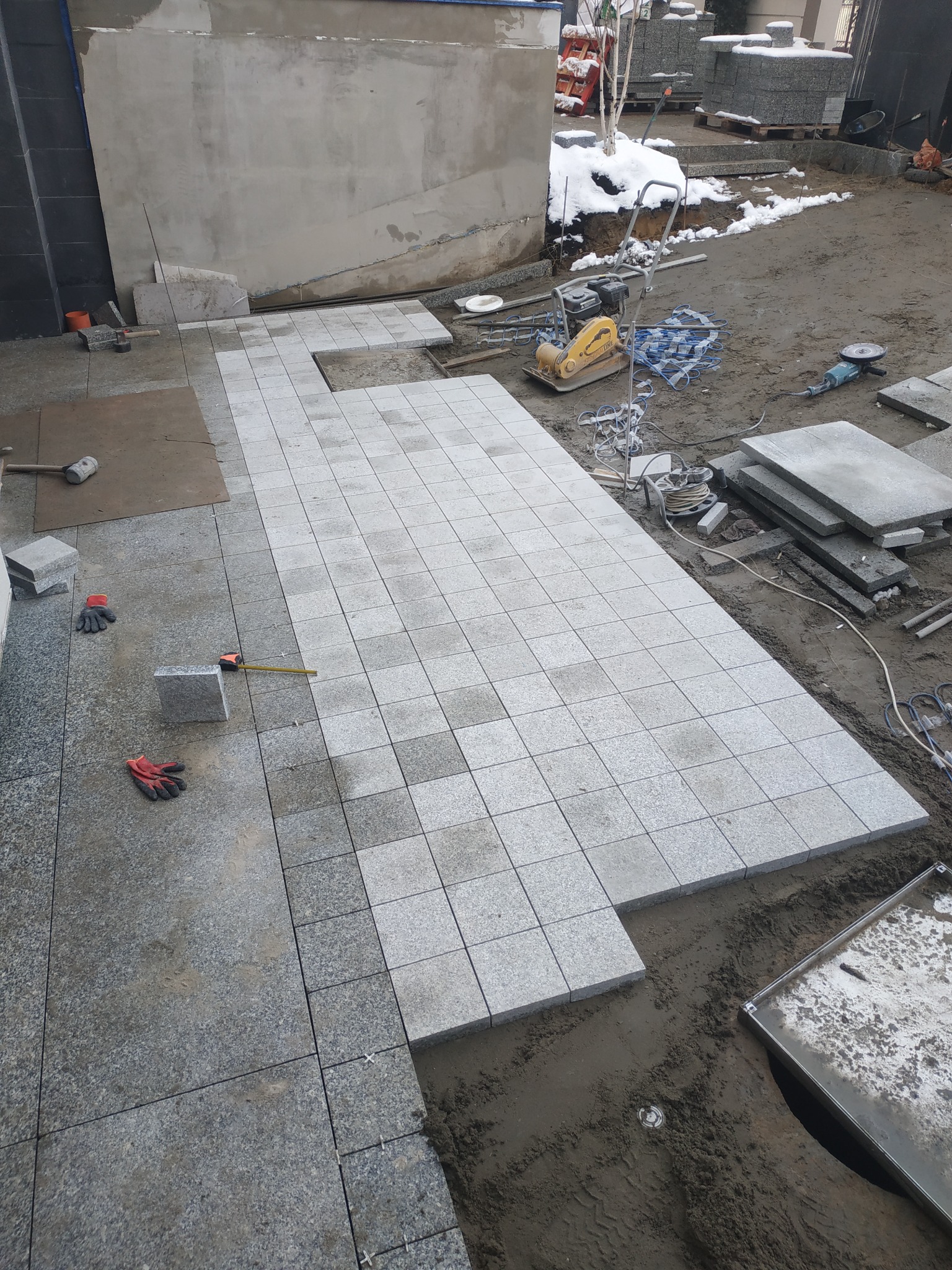 Układanie jasnoszarej kostki na placu budowy, widoczna ubijarka, szlifierka kątowa, palety z kostką w tle i resztki śniegu.