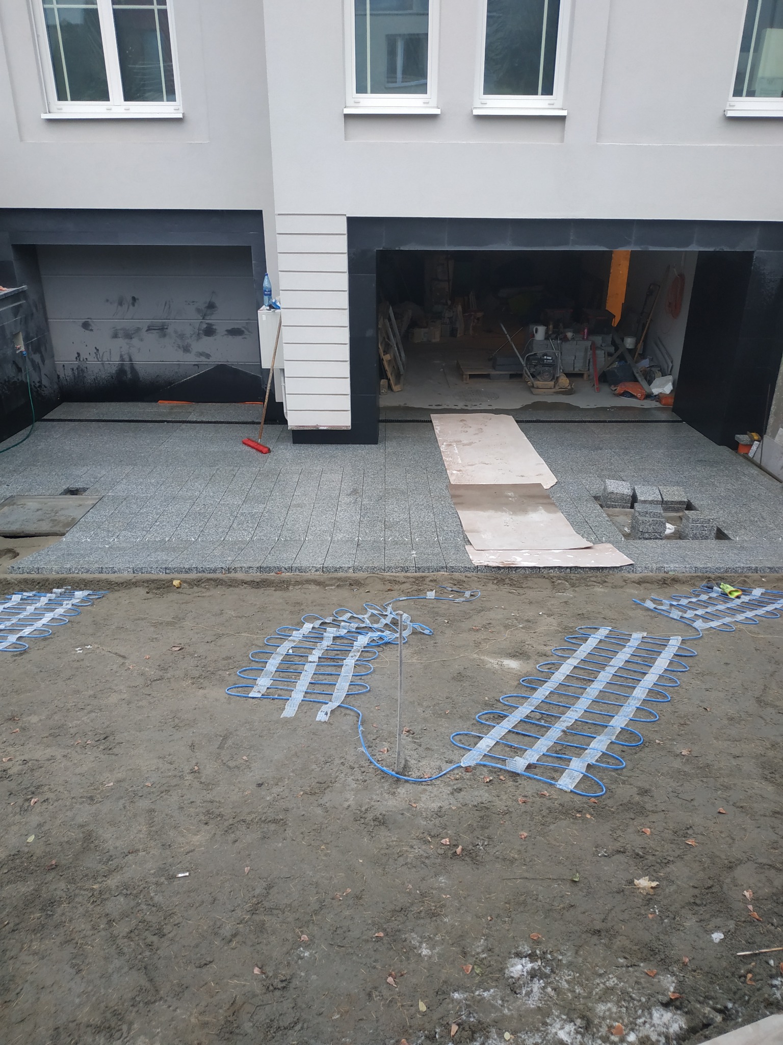 Montaż ogrzewania podjazdowego z kostki granitowej: widoczne maty grzewcze na ziemi przed częściowo ułożonym podjazdem z kostki granitowej w kolorze szarym, garaż w tle.