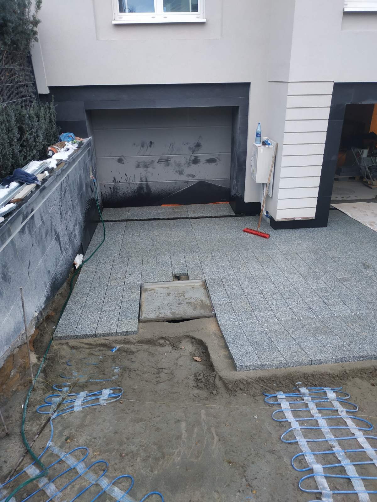 Układanie szarej kostki granitowej na podjeździe, widoczne kable grzewcze w piasku, studzienka rewizyjna i częściowo ułożona powierzchnia przed garażem.