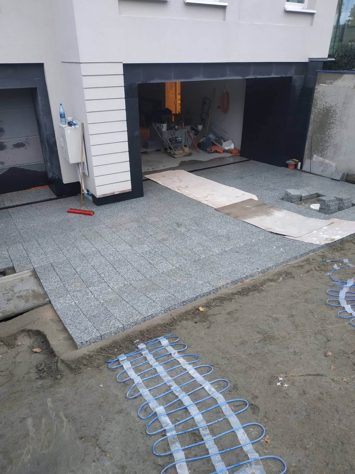Fragment podjazdu z ułożoną kostką granitową na siatce, widoczne kable grzewcze w ziemi, w tle garaż i narzędzia brukarskie.