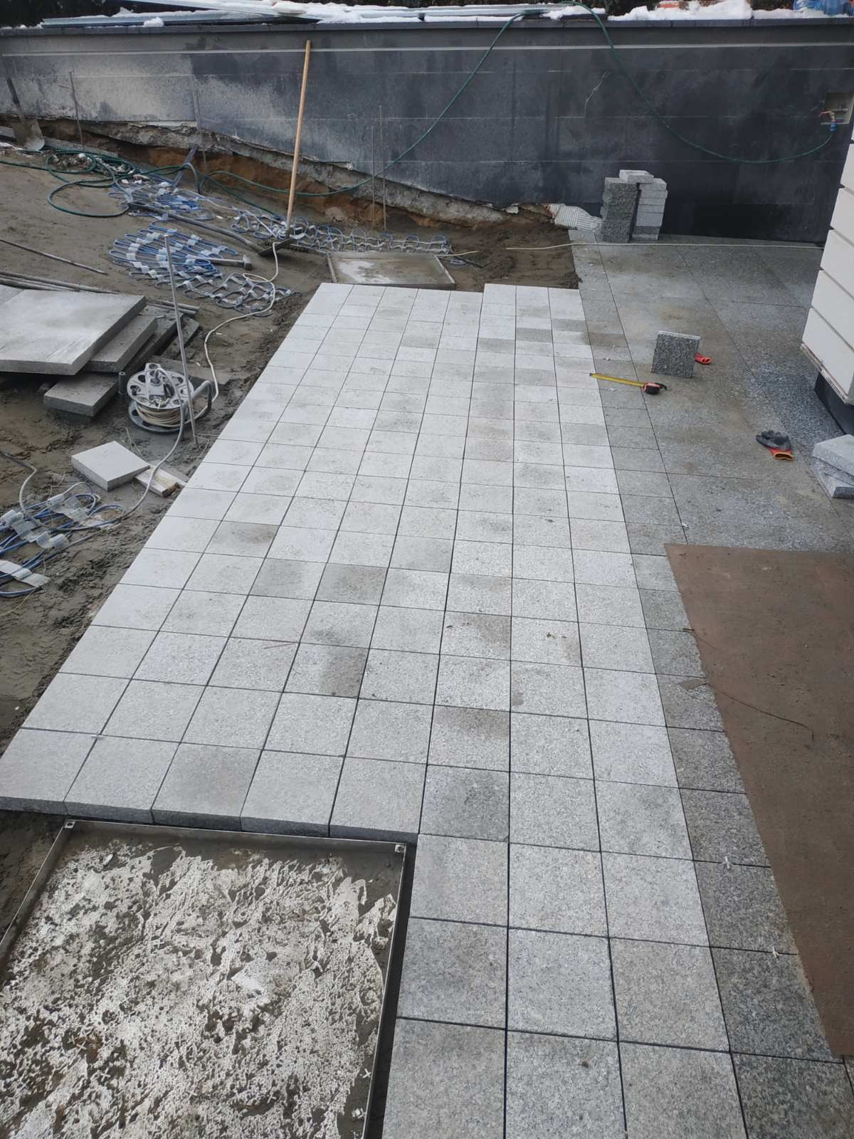 Układanie jasnoszarej kostki granitowej w kwadratowy wzór, widoczne narzędzia, częściowo ułożona nawierzchnia i przygotowane podłoże z przewodami grzewczymi.