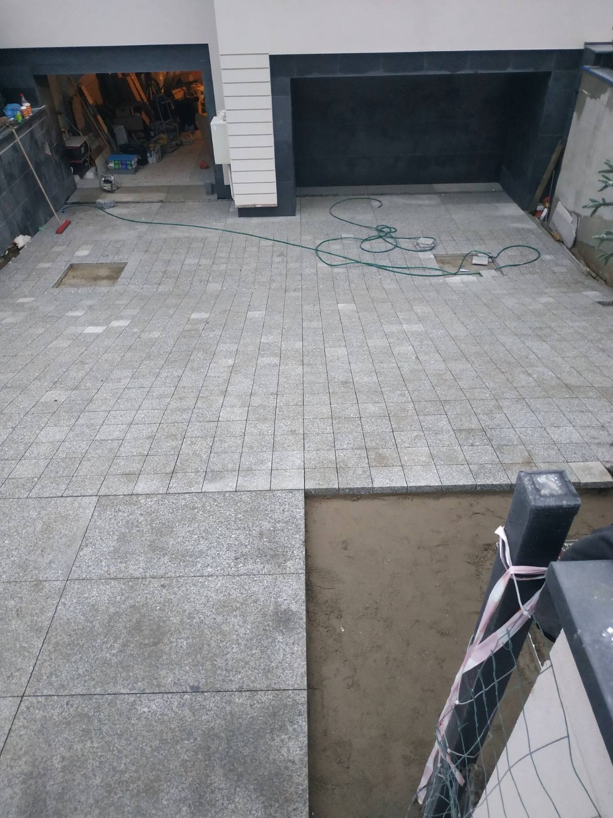Fragment układanej nawierzchni z jasnoszarej kostki granitowej, widoczne niedokończone fragmenty, węże ogrodowe i czerwona zmiotka w tle.