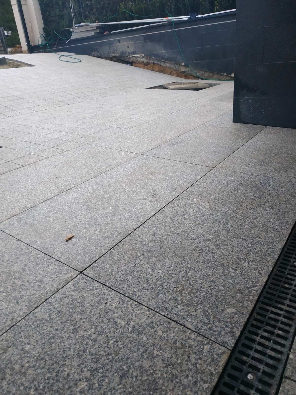 Ułożona szara kostka granitowa na zewnątrz budynku, z widoczną kratką odpływową i małym liściem na powierzchni.