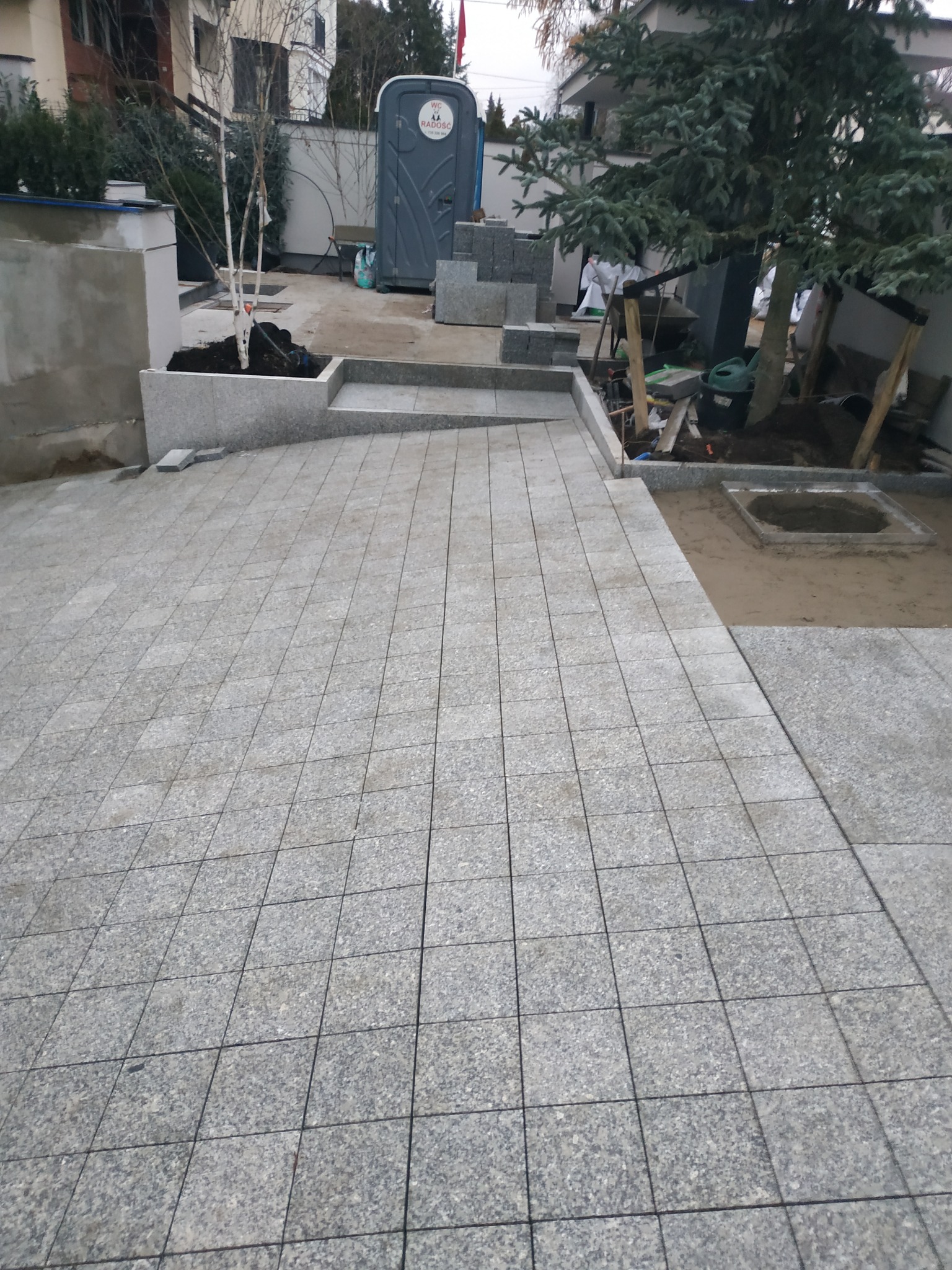 Ułożona szara kostka granitowa na podwórku z elementami ogrodu i tymczasową toaletą w tle.