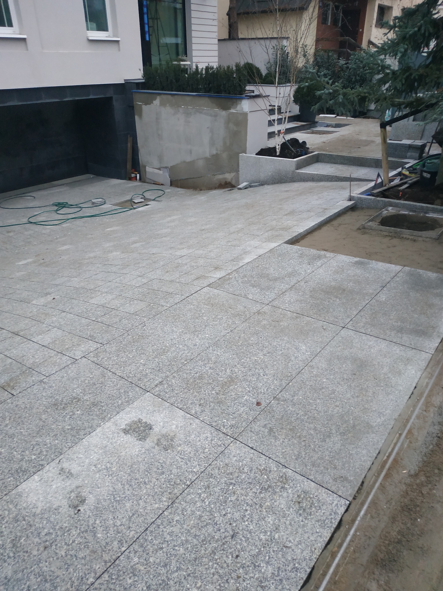 Fragment układanej szarej kostki granitowej na podjeździe, widoczne narzędzia i elementy ogrodu w trakcie tworzenia.