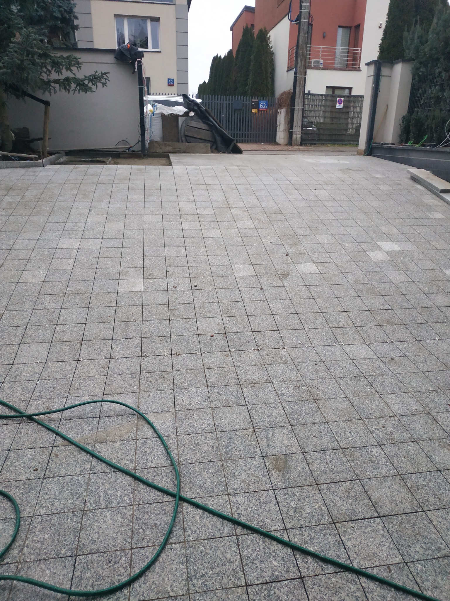 Ułożona szara kostka granitowa na podjeździe domu jednorodzinnego, widoczny zielony wąż ogrodowy i przygotowane podłoże pod dalszą część nawierzchni.