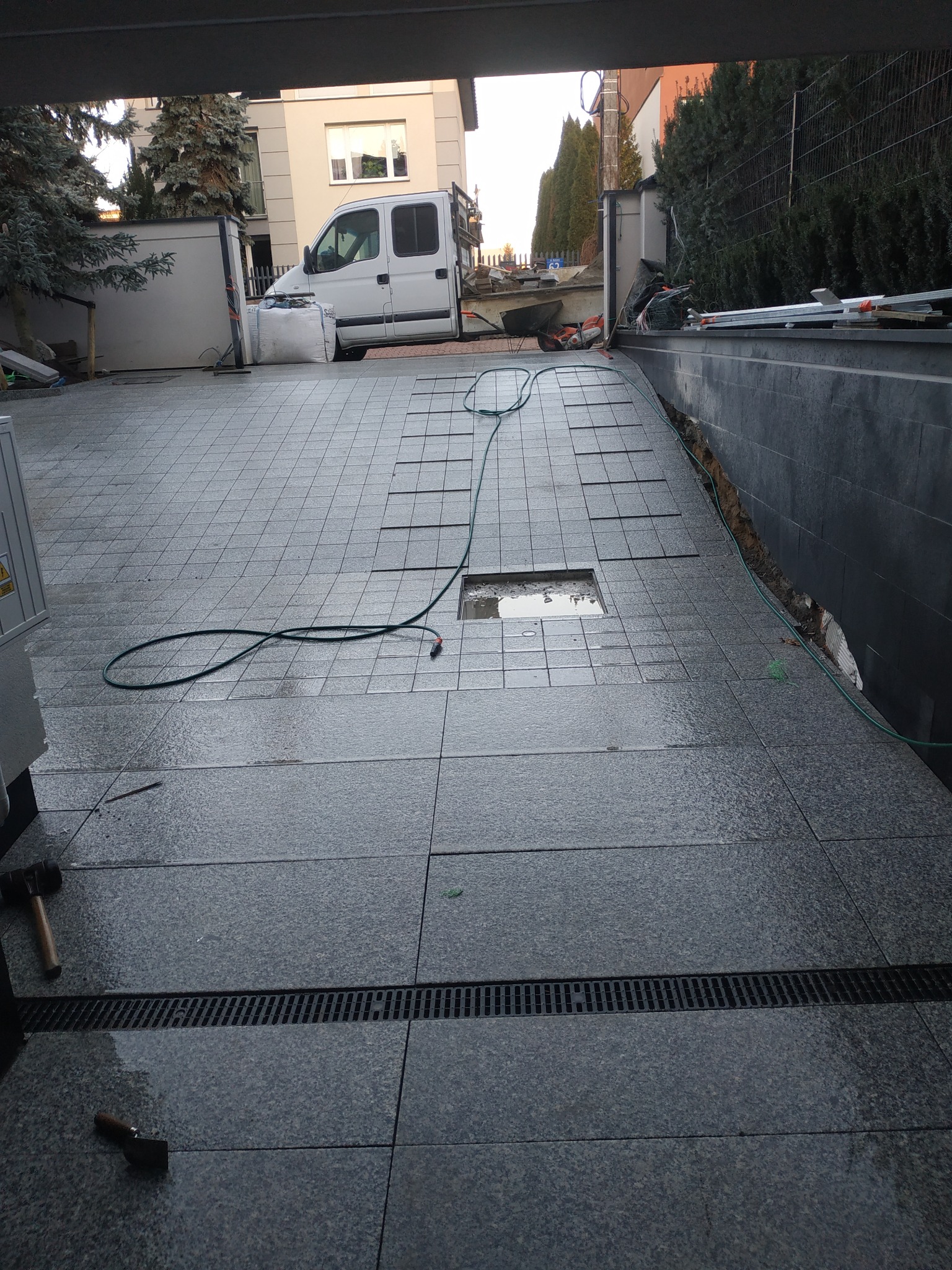 Położona częściowo granitowa nawierzchnia w trakcie prac, widoczny fragment z kratką odpływową, wąż ogrodowy oraz narzędzia brukarskie, w tle samochód dostawczy i budynek mieszkalny.