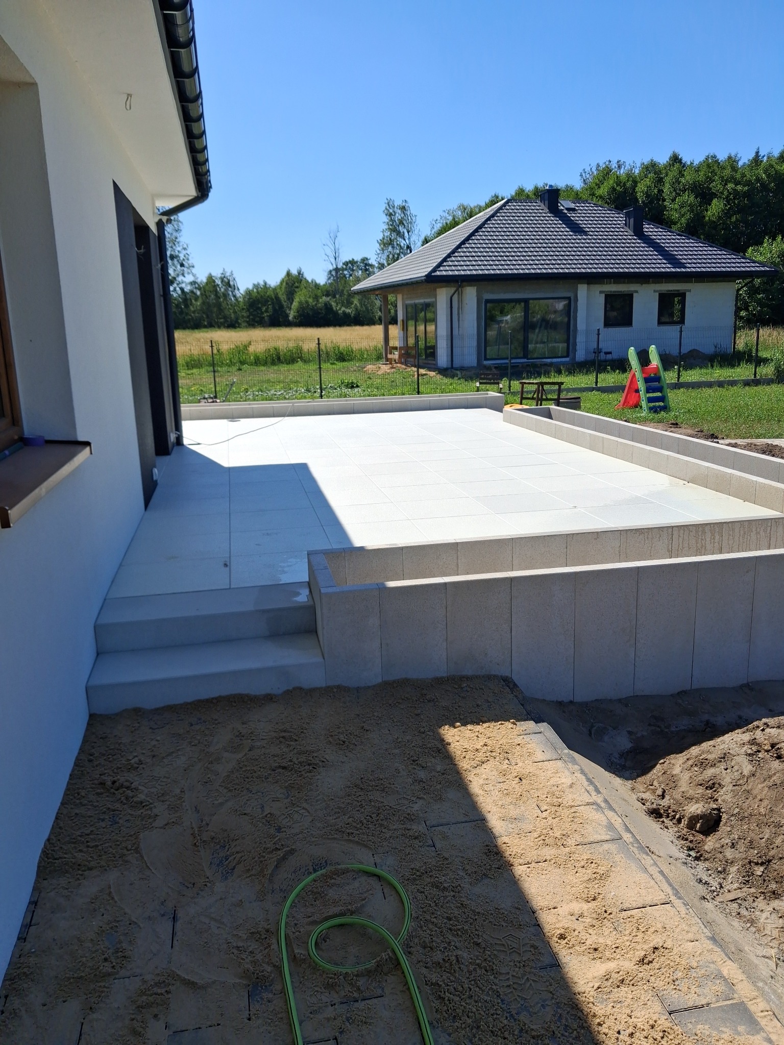 Nowoczesny taras z jasnych płyt, obrzeża z kostki, w tle dom jednorodzinny i zieleń. Widoczny fragment przygotowanego podłoża pod dalszą część tarasu.