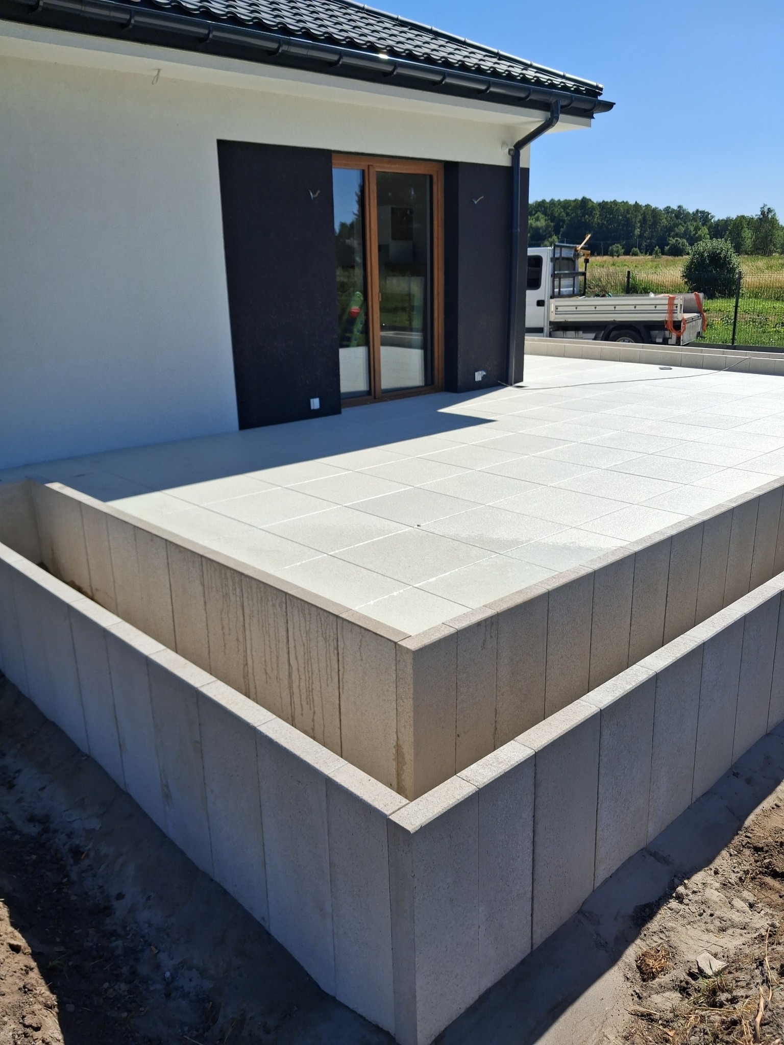 Jasne, kwadratowe płyty tarasowe ułożone na podwyższeniu z betonowych elementów. Nowoczesny dom z przesuwnymi drzwiami w tle, obok zaparkowany biały samochód dostawczy.