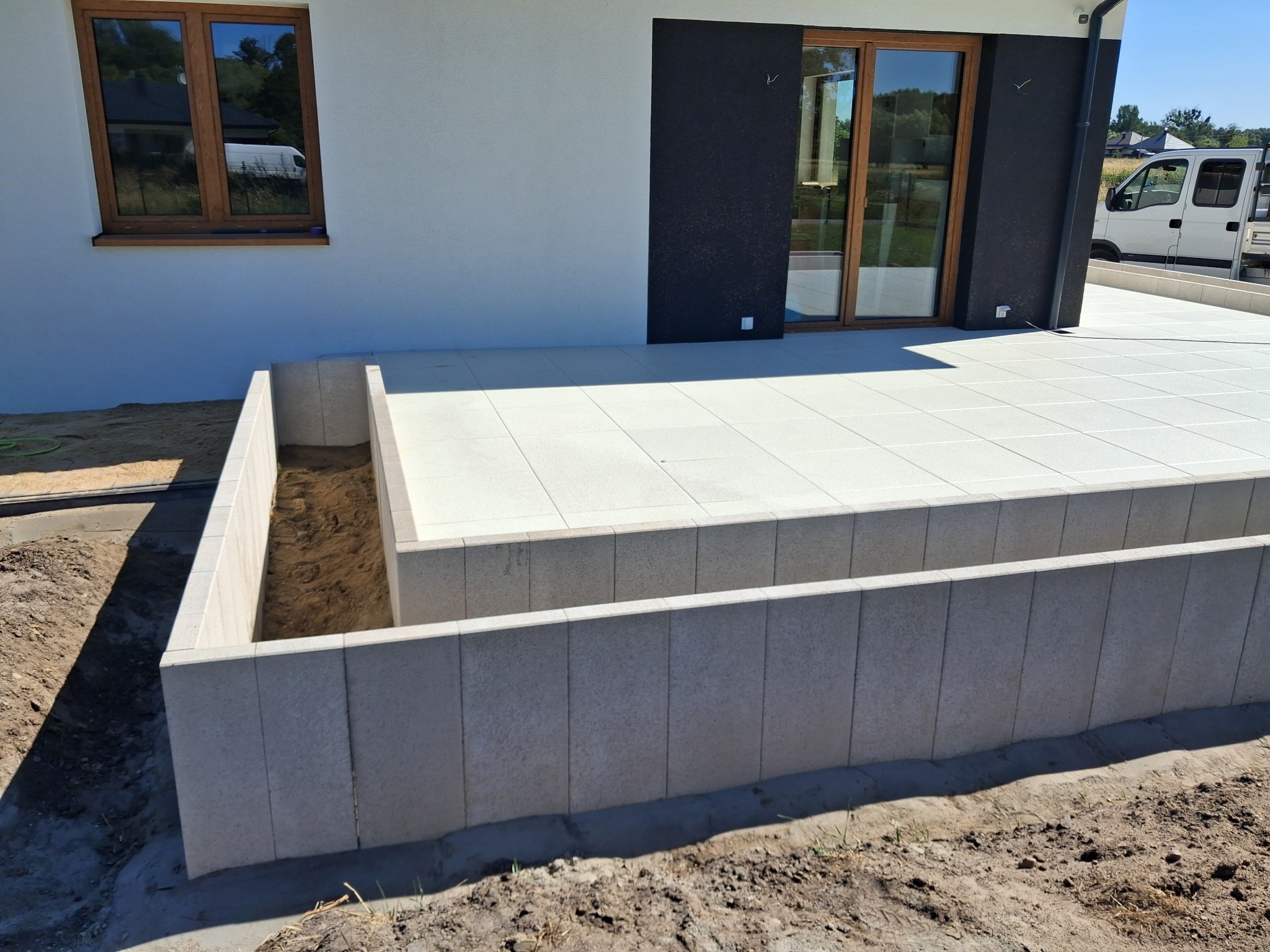 Nowoczesny taras z jasnych płyt na zewnątrz domu z antracytowym akcentem. Widoczna obudowa rabaty z kostki. W tle zaparkowany biały bus.