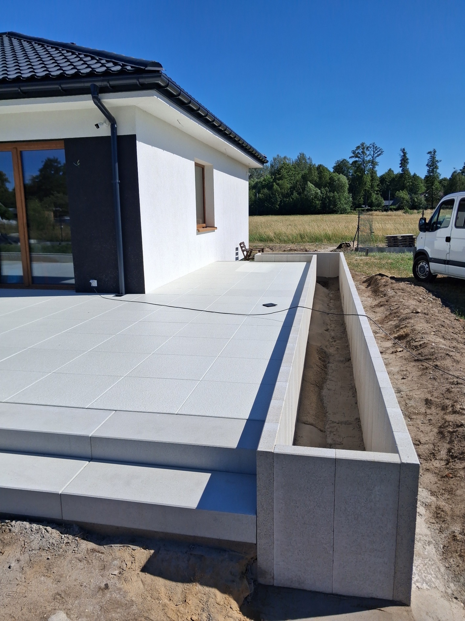 Jasnoszara, nowoczesna kostka brukowa tworząca taras przy domu z widokiem na pole. Widoczny fragment wkopanej donicy i samochód dostawczy.