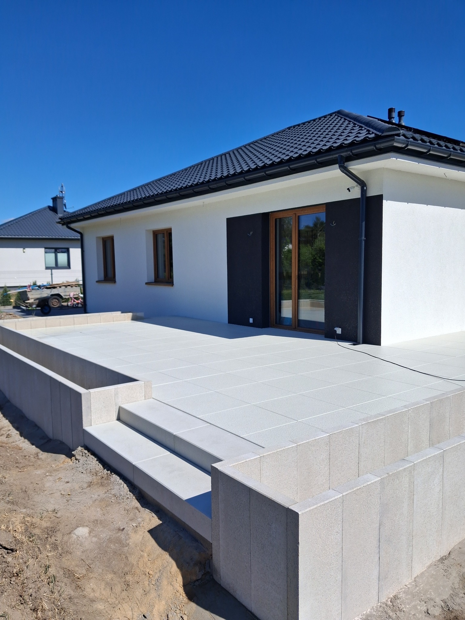 Jasnoszara, ułożona kostka brukowa tworzy taras przed nowoczesnym domem. Widoczne schody z kostki i fragment ogrodu. Słoneczny dzień, błękitne niebo.