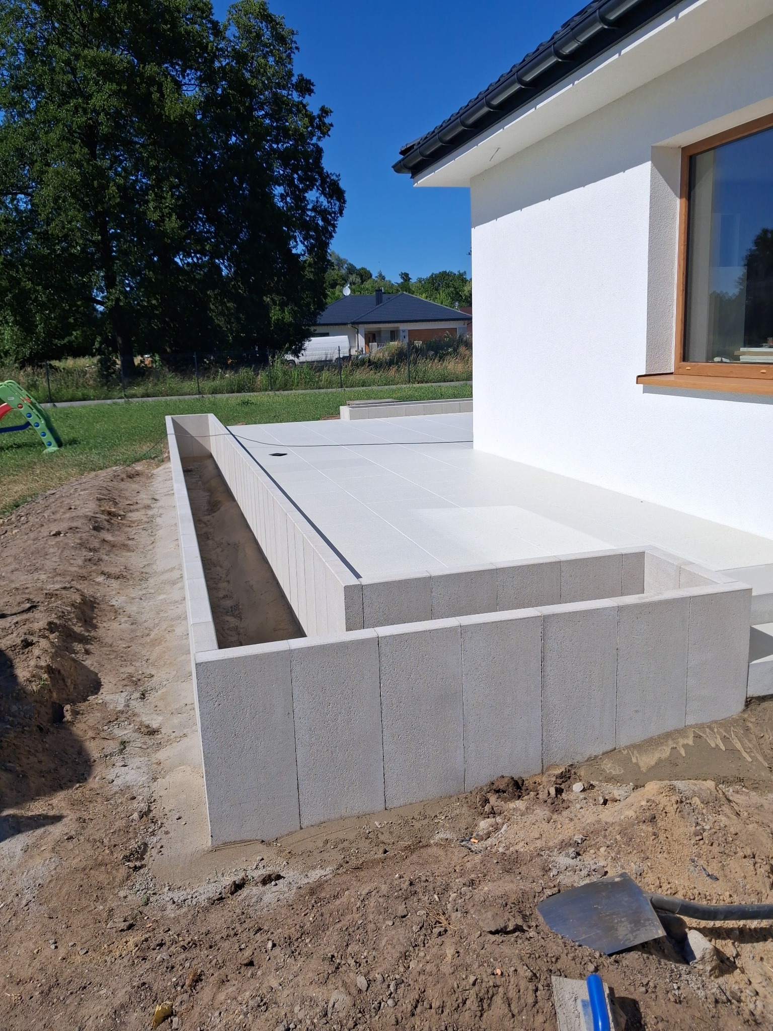 Białe płyty tarasowe obramowane betonowymi bloczkami przy ścianie domu. Widoczny fragment ogrodu i niebo. Szpadel na ziemi sugeruje prace wykończeniowe.