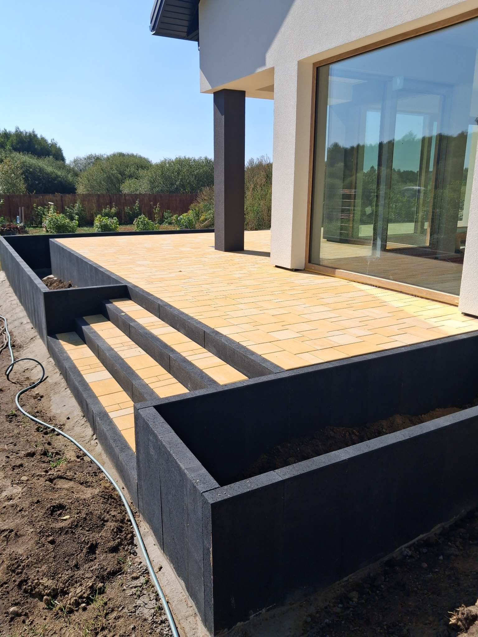 Żółta kostka brukowa na tarasie z czarnymi obrzeżami i schodami, w tle nowoczesny dom z dużym oknem, widok z dołu, słoneczny dzień.