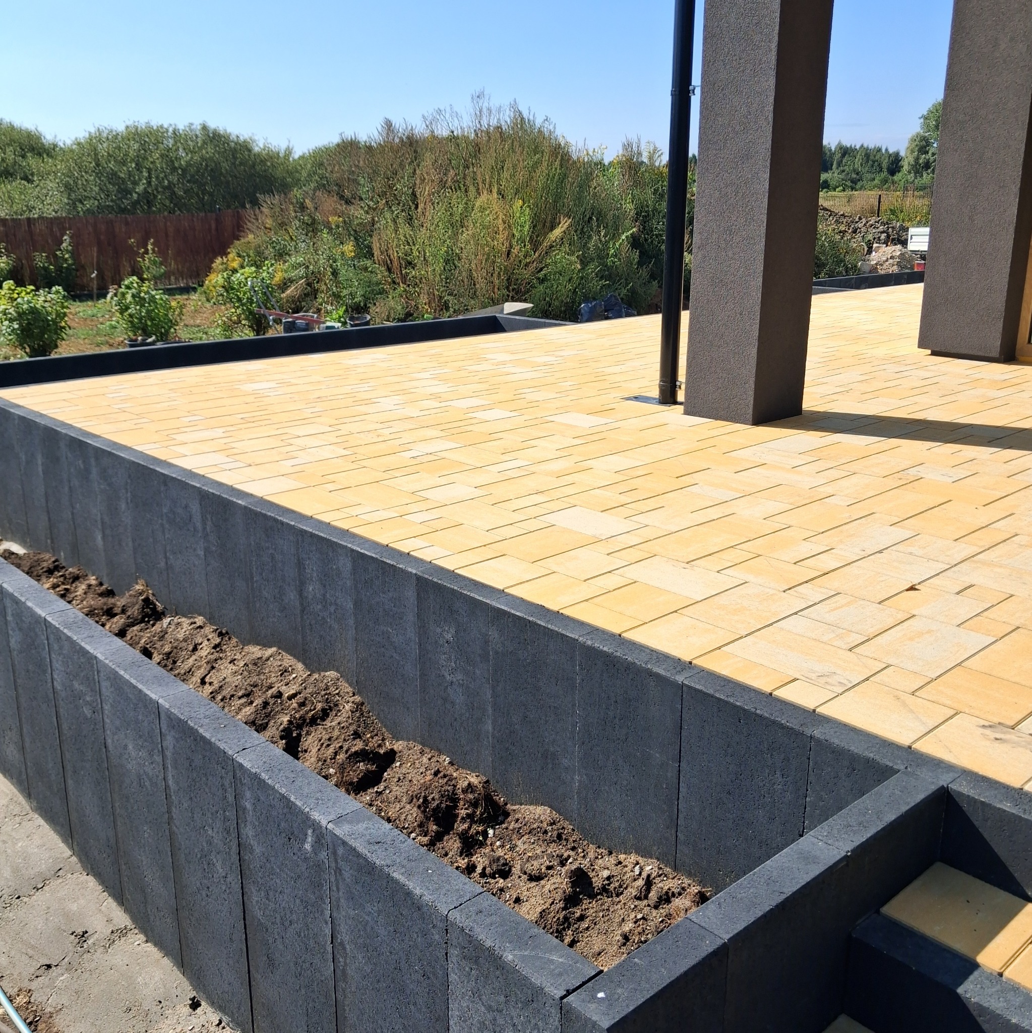 Żółta kostka brukowa ułożona na tarasie z donicą z szarej kostki. Widok na zieleń w tle. Słoneczny dzień.