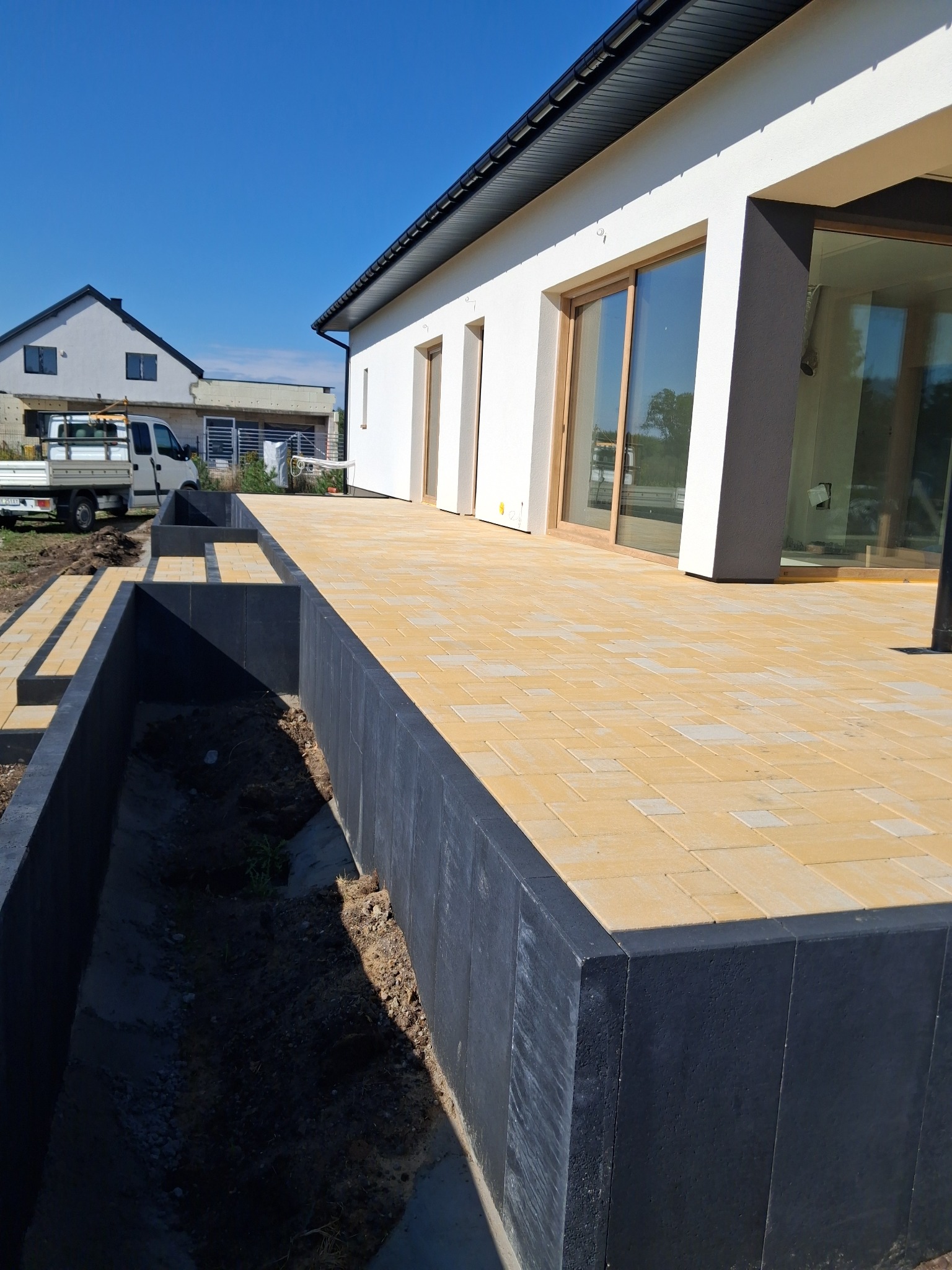 Żółta kostka brukowa na tarasie nowoczesnego domu z szarą podmurówką, widok z dołu na błękitne niebo w słoneczny dzień. W tle biały budynek i samochód dostawczy.