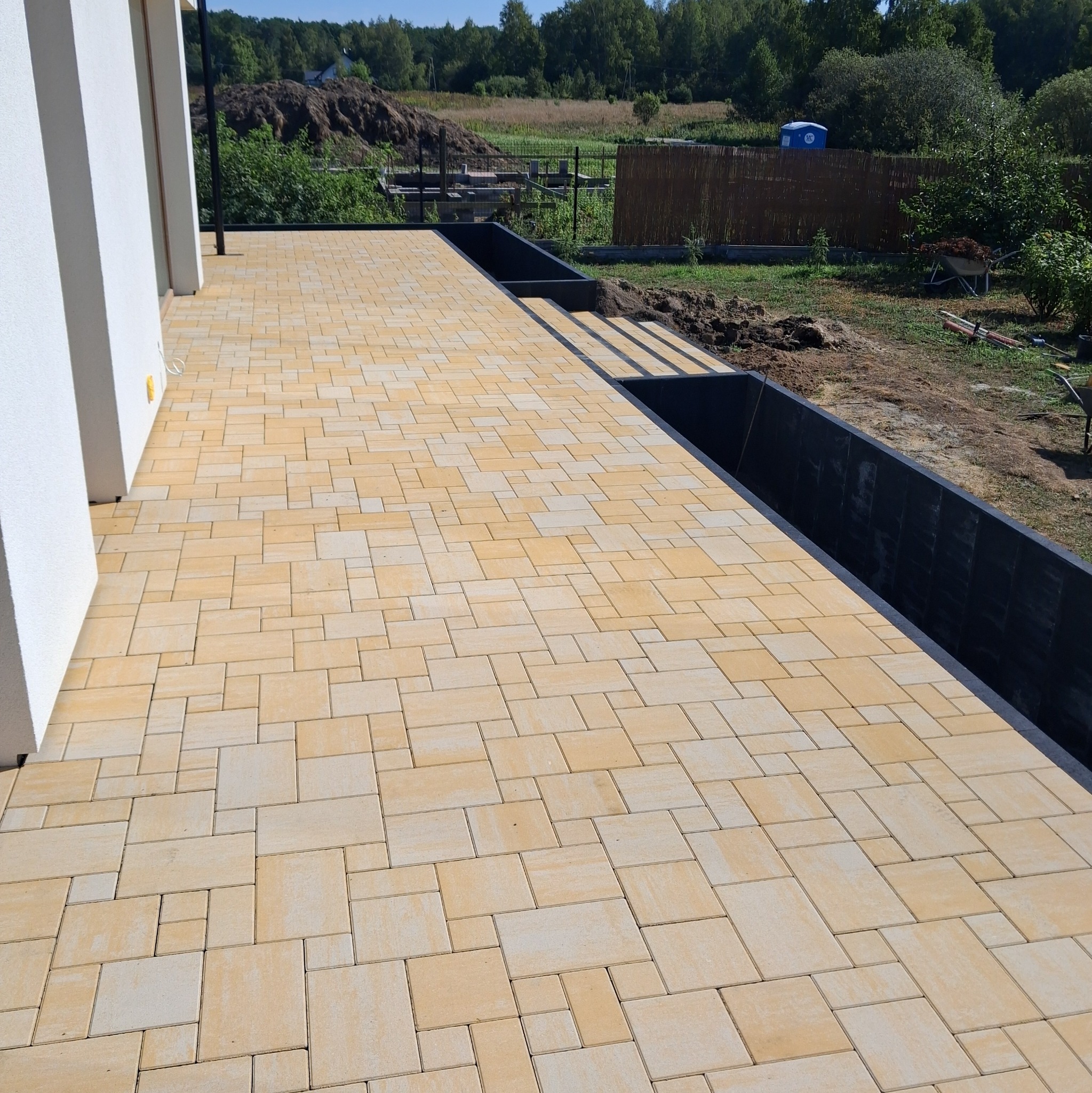 Jasna kostka brukowa ułożona w nieregularny wzór na tarasie domu, z widokiem na zielony ogród i elementy konstrukcyjne w tle. Nowoczesne wykończenie.