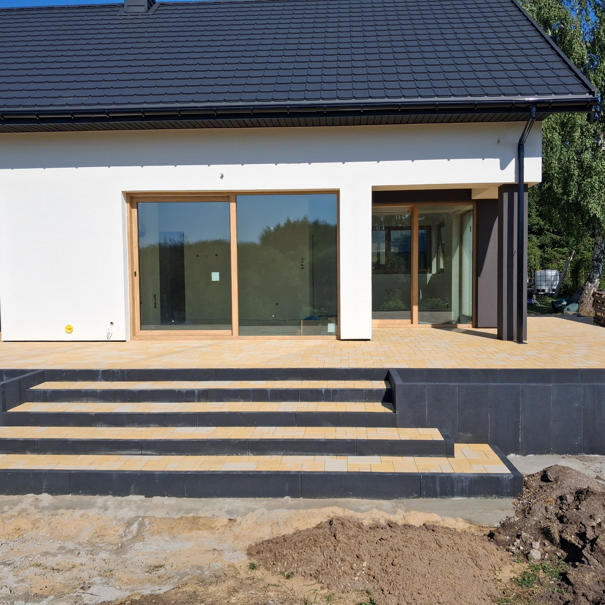 Nowoczesny taras z kostki brukowej w odcieniach beżu i szarości, prowadzący do domu z dużymi oknami przesuwnymi. Widoczne schody i fragment trawnika.