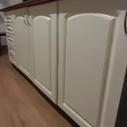 Fragment zabudowy kuchennej z kremowymi, frezowanymi frontami szafek i szuflad, zwieńczonej drewnianym blatem, widzianej z niskiej perspektywy na tle drewnianej podłogi.