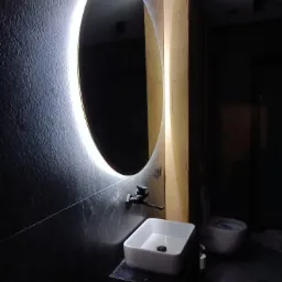 Nowoczesna łazienka z podświetlanym, okrągłym lustrem, czarną armaturą i kwadratową umywalką na ciemnym blacie, widoczna toaleta w tle.