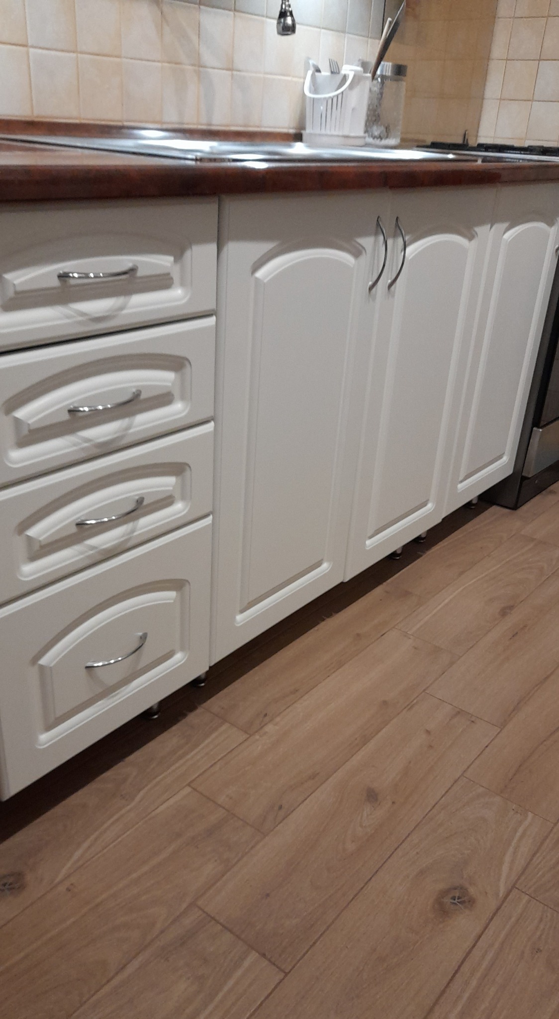 Białe szafki kuchenne z frezowanymi frontami i chromowanymi uchwytami, brązowy blat, jasnobrązowa podłoga z paneli.