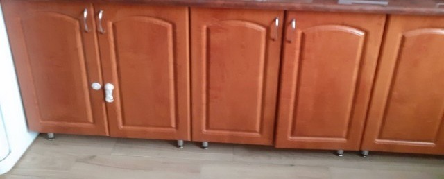 Dolne szafki kuchenne z brązowego drewna z metalowymi uchwytami i małymi nóżkami, widoczne na tle jasnej podłogi.