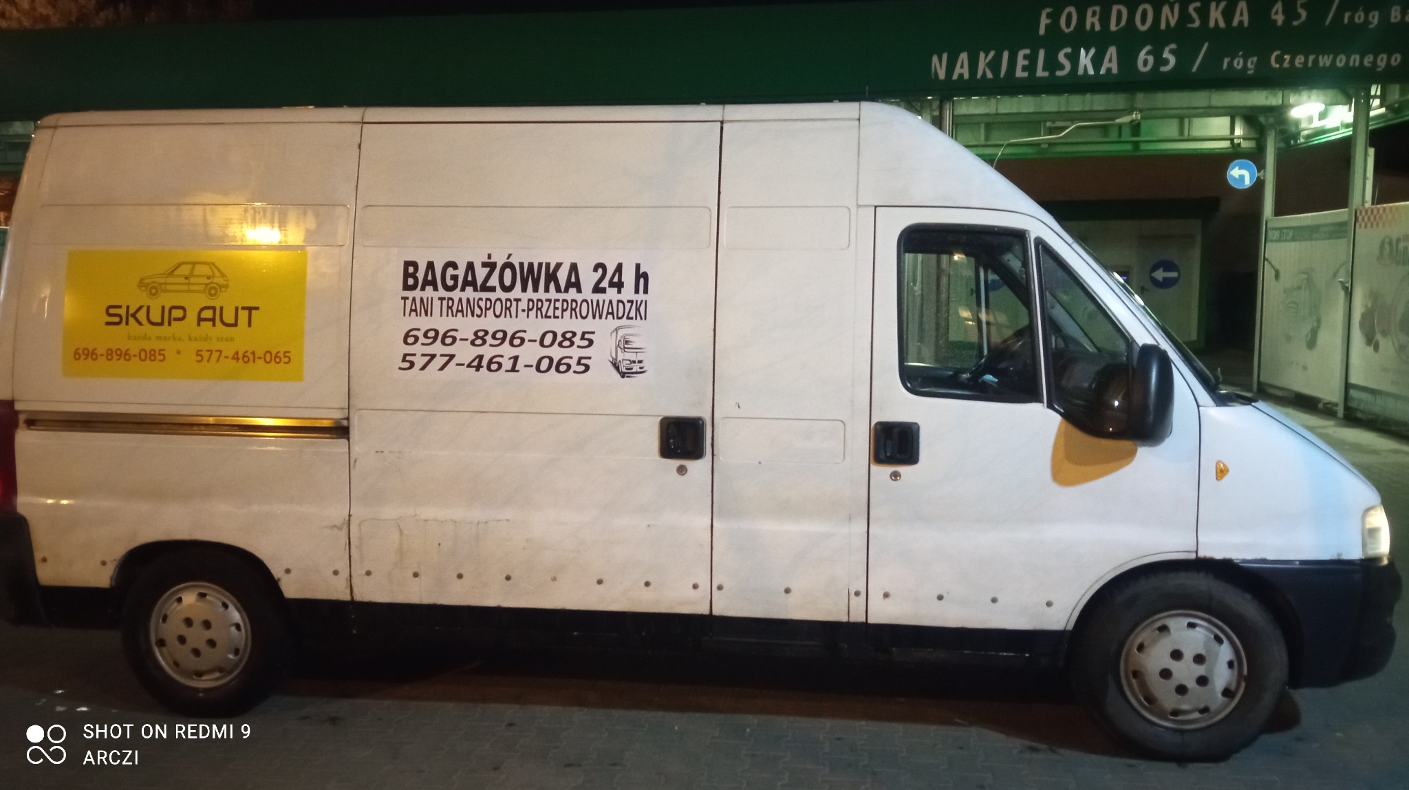 Biały bus dostawczy z reklamą usług transportowych i przeprowadzkowych, zaparkowany nocą przy ulicy.