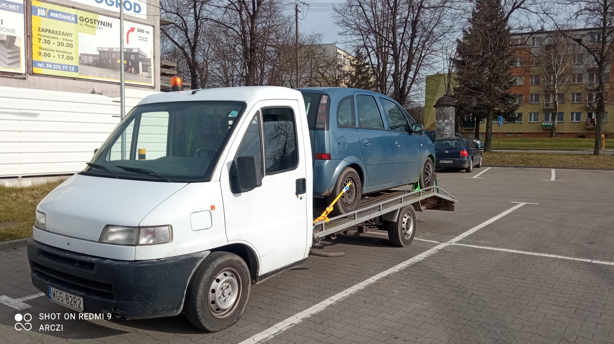 Biała laweta z niebieskim samochodem osobowym Opel Zafira na platformie, zabezpieczonym pasami transportowymi, zaparkowana na parkingu z widocznymi budynkami mieszkalnymi i reklamą sklepu...