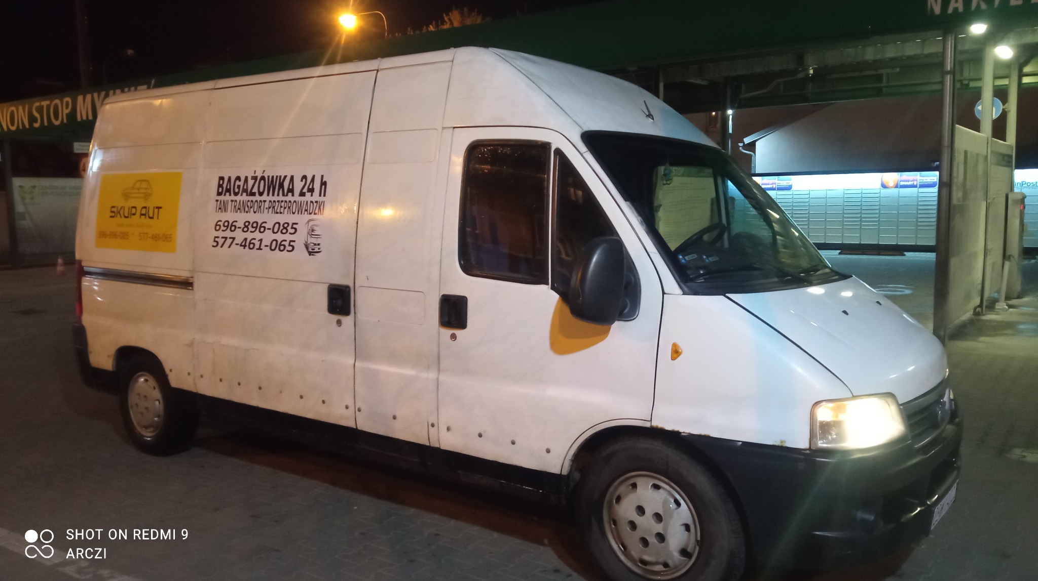 Biały samochód dostawczy z napisem 'Bagażówka 24h Tani Transport-Przeprowadzki' i numerami telefonu, zaparkowany w nocy przy paczkomacie.