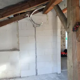 Instalacja przewodów wentylacyjnych i elektrycznych w surowym pomieszczeniu z bloków betonowych, widoczne drewniane elementy konstrukcyjne dachu, skrzynka narzędziowa w tle.