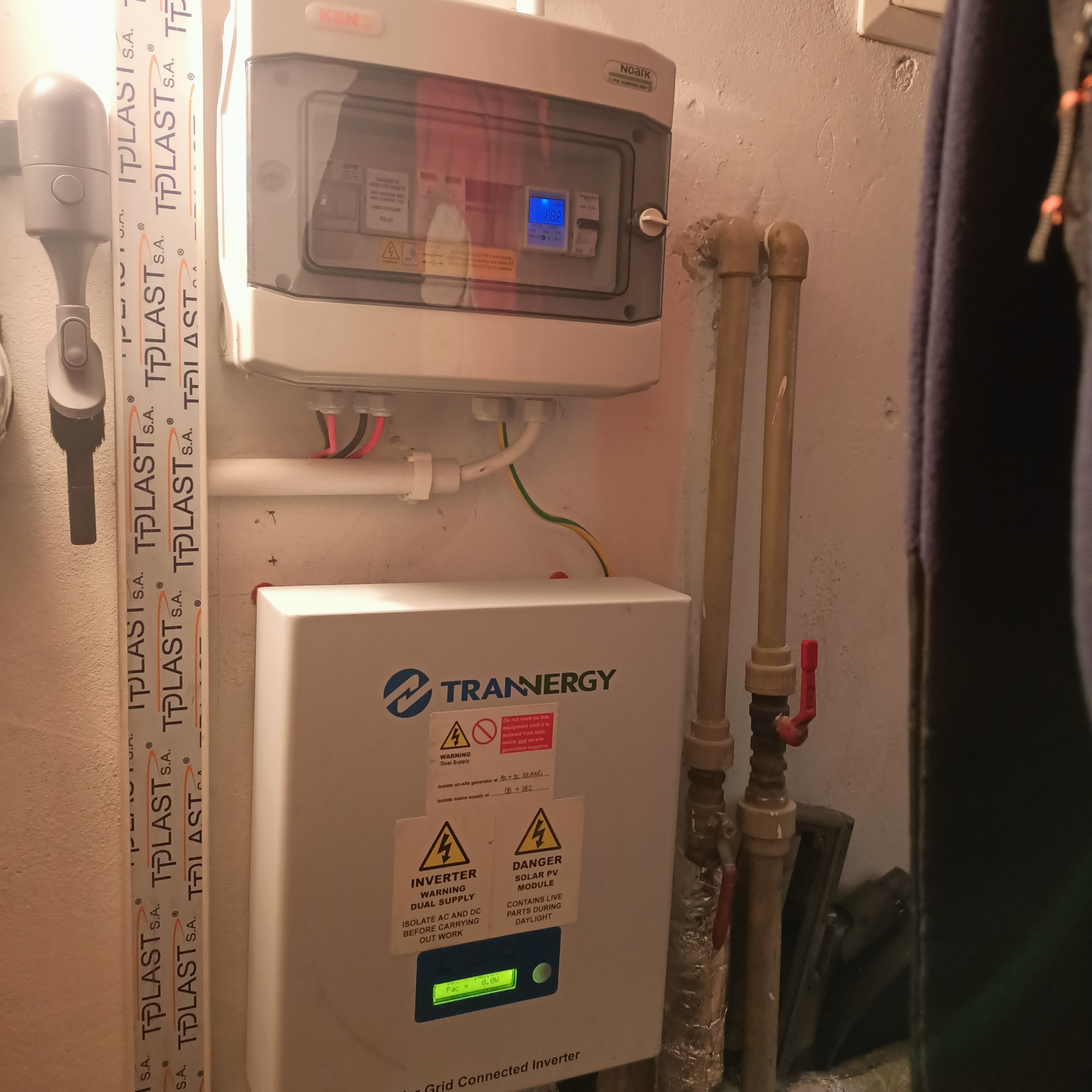 Biała skrzynka elektryczna z wyświetlaczem i oznaczeniami ostrzegawczymi, zamontowana na ścianie obok rur, z widocznym inwerterem i zabezpieczeniami.