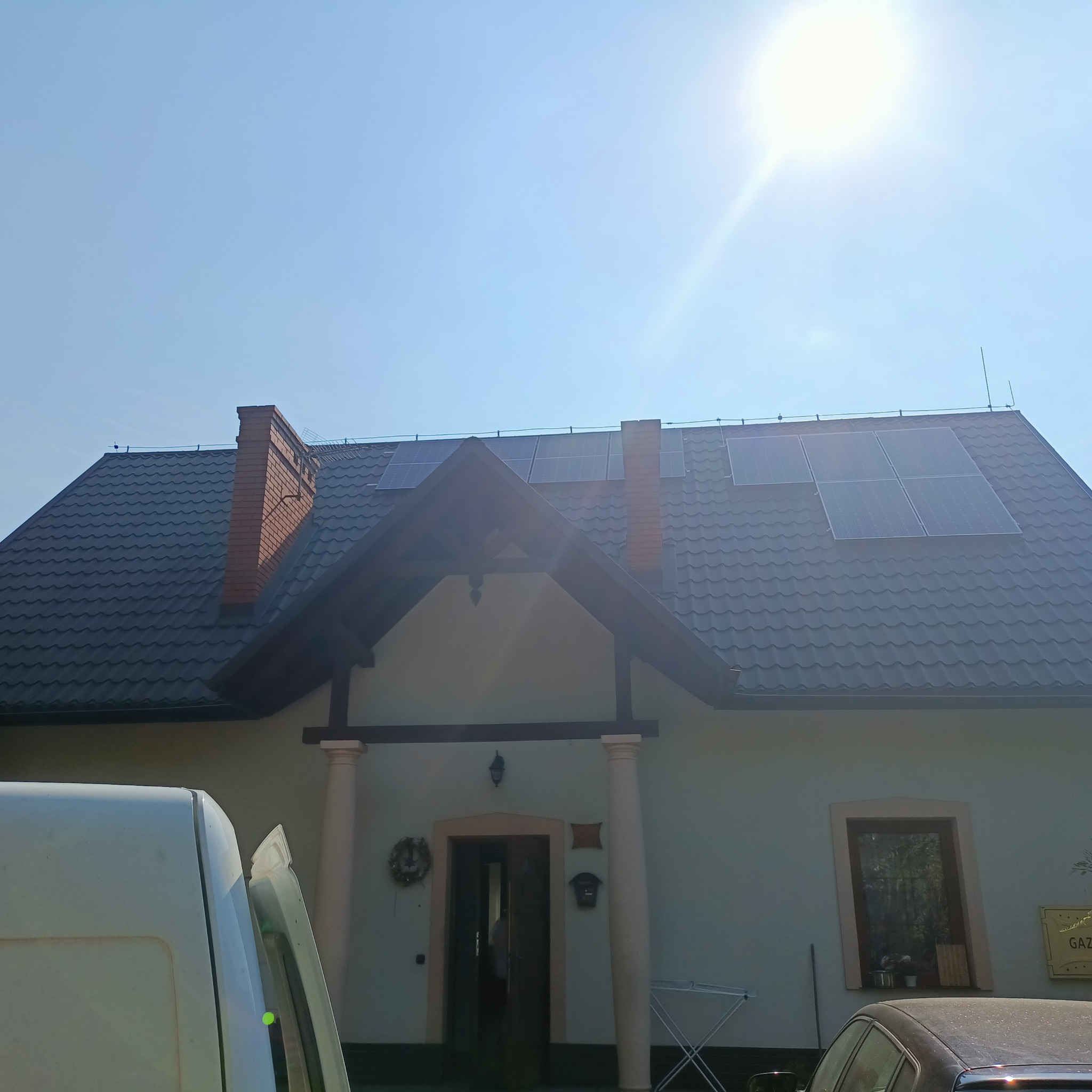 Dom jednorodzinny z ciemną dachówką i panelami słonecznymi, częściowo zasłonięty przez biały samochód dostawczy i czarny samochód osobowy, widok w słoneczny dzień.