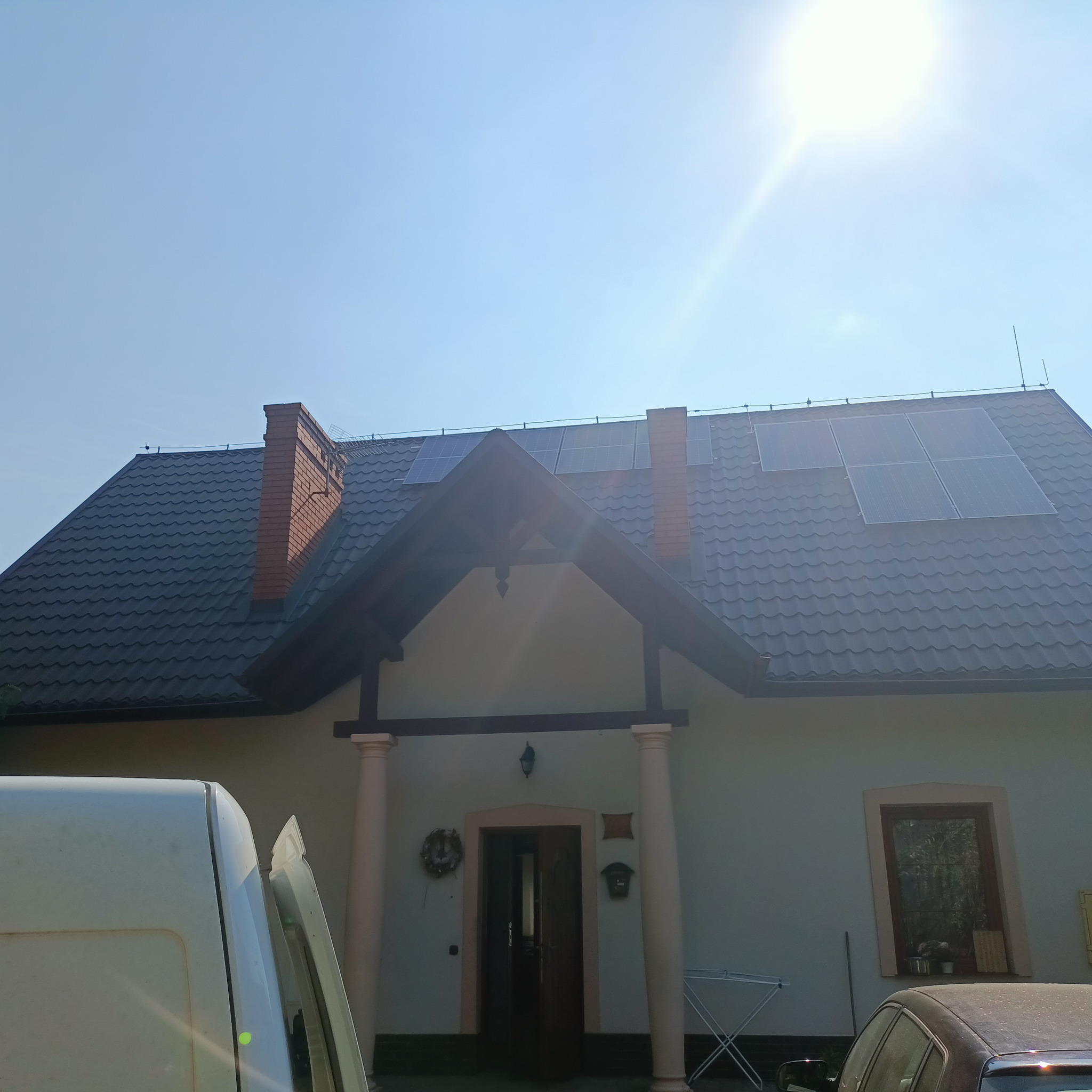 Dom jednorodzinny z zainstalowanymi panelami fotowoltaicznymi na dachu pokrytym ciemną dachówką, widoczne także kominy z czerwonej cegły, fragment samochodu i busa na pierwszym planie.