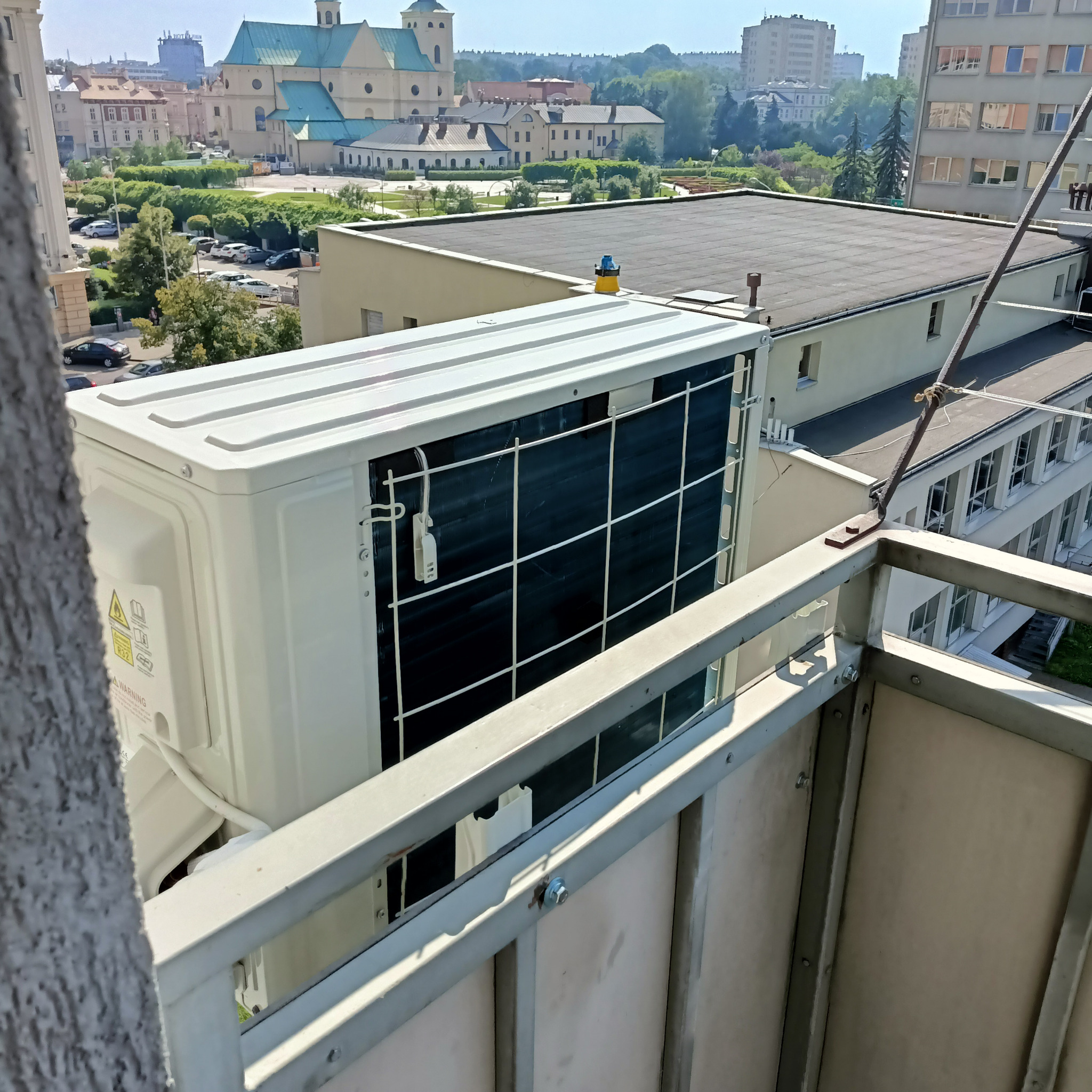 Biała jednostka zewnętrzna klimatyzatora zamontowana na balkonie, widok na panoramę miasta z kościołem w tle.