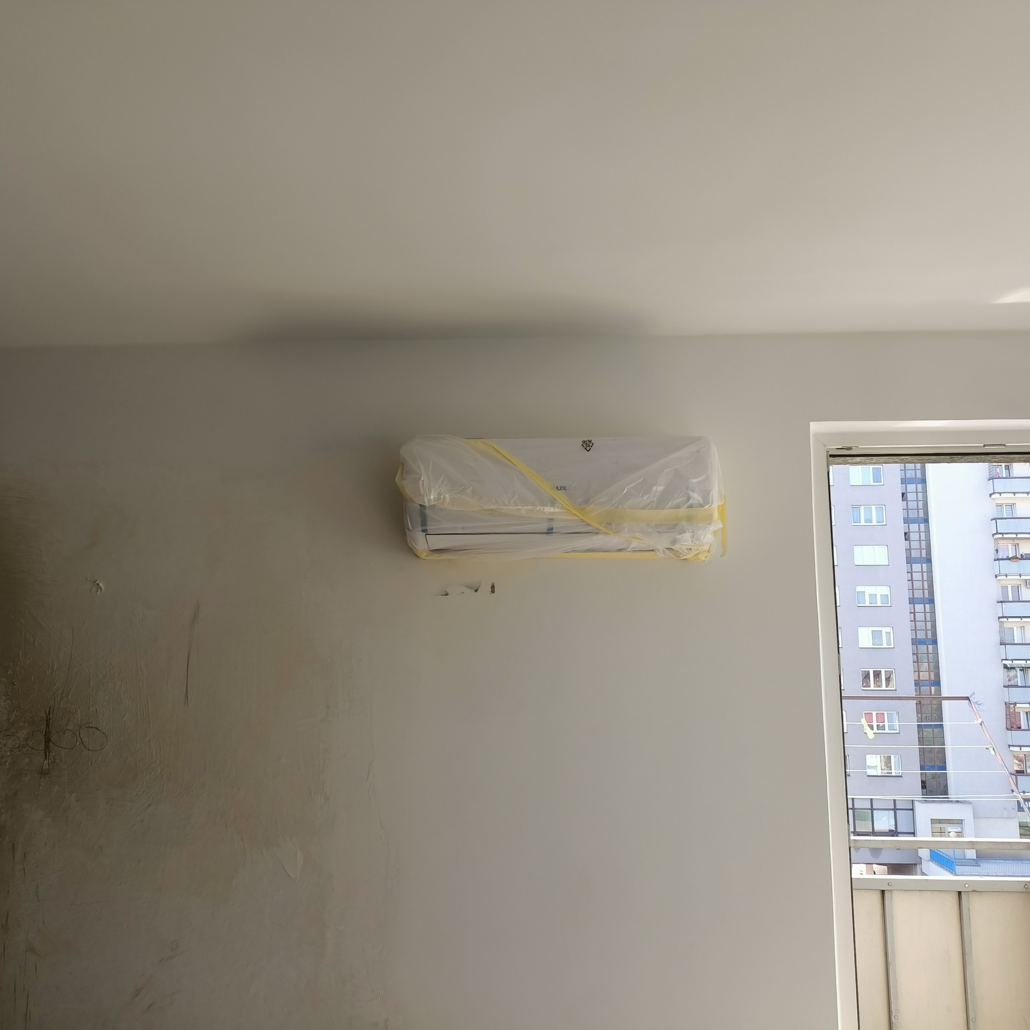 Nowy klimatyzator zabezpieczony folią i taśmą malarską na ścianie w trakcie remontu mieszkania, widok przez okno na blok mieszkalny.